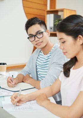 Modern Asian man in glasses looking at camera while sitting with notepad at table wit female colleague